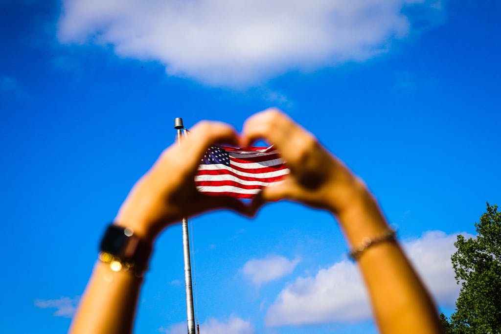 Hands form a heart shape around an American flag against a vibrant blue sky, symbolizing patriotism and freedom.